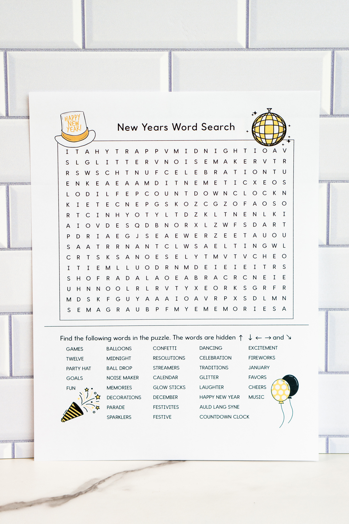 Alt text: A New Year's word search printable displayed against white subway tile, featuring a large word search grid with small festive illustrations like a party hat, disco ball, and balloons, along with a list of themed words to find.