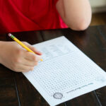 Alt text: A child wearing a red shirt sits at a dark wooden table, holding a yellow pencil while working on a New Year’s word search printable, with the puzzle and word list visible.
