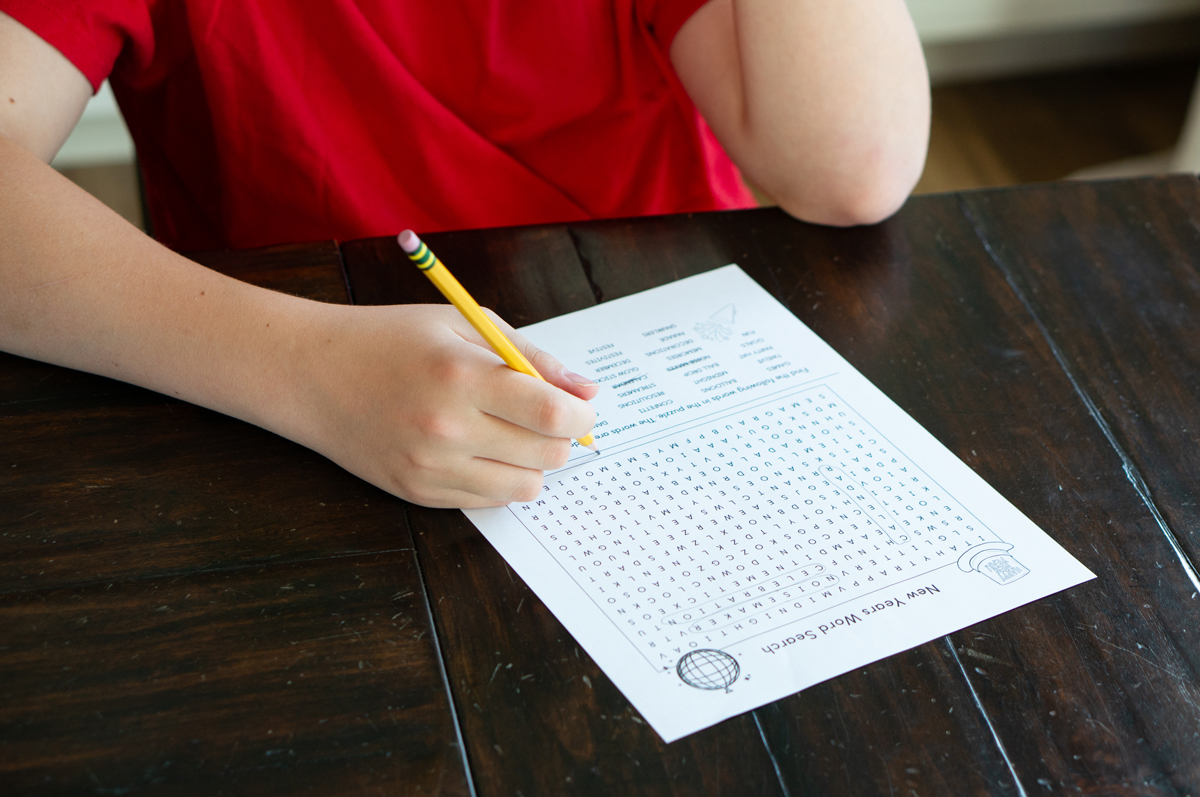 Alt text: A child in a red shirt works on a New Year's word search printable at a dark wooden table, holding a yellow pencil above the puzzle grid and word list.