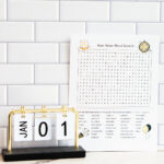 Alt text: A New Year's word search printable propped beside a small desktop flip calendar showing “JAN 01,” set against a white subway tile background; the printable includes a festive word search and themed illustrations.