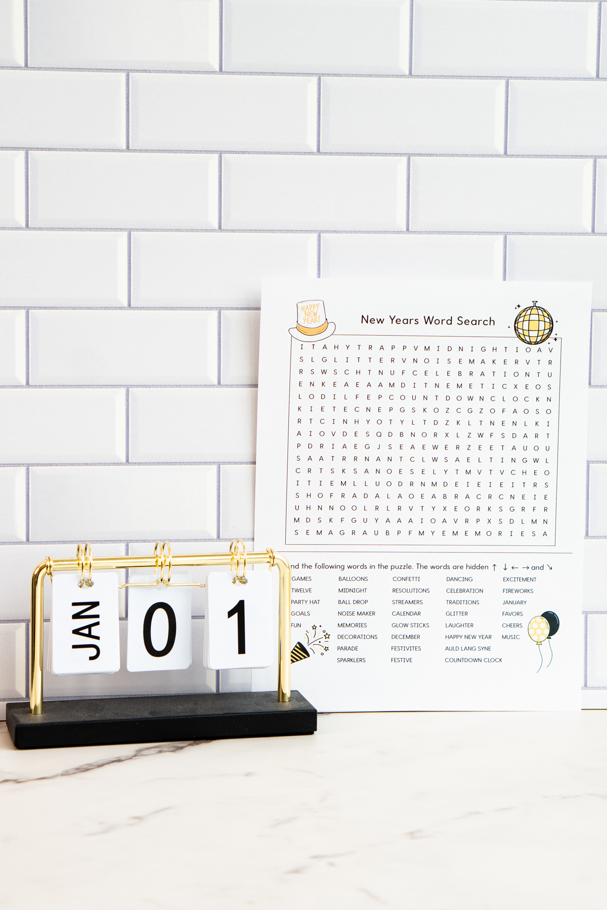 Alt text: A New Year's word search printable propped beside a small desktop flip calendar showing “JAN 01,” set against a white subway tile background; the printable includes a festive word search and themed illustrations.