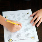 Alt text: A child working on the New Year’s word search at a dark wooden table, holding a yellow pencil and circling words on the printable decorated with festive illustrations.
