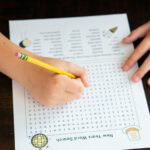 Alt text: Close-up of a child’s hands using a yellow pencil to work on a New Year’s word search printable at a dark wooden table, showing part of the themed word list and illustrations.