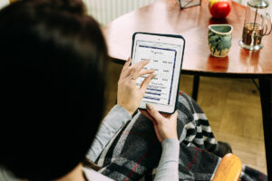 Woman sitting in a cozy chair with a blanket, holding a tablet that displays the digital planner&rsquo;s templates page, showing options like lined, dot grid, graph, and blank layouts.