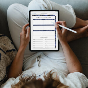 Person sitting comfortably with an iPad on their lap, filling out the February Overview page of the digital planner using a stylus, with sections for accomplishments, challenges, and &ldquo;Currently&rdquo; prompts.