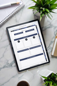 Flat-lay image of an iPad on a marble desk showing the Goal 1 page of the digital planner, surrounded by a coffee cup, notebook, pencil, and small potted plants.