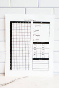 Yearly period tracker printable displayed against a white subway tile backdrop. The page features a 31-day vertical grid labeled with months, a simple flow-level key for spotting through heavy, a monthly cycle length chart, and a lined notes section at the bottom.