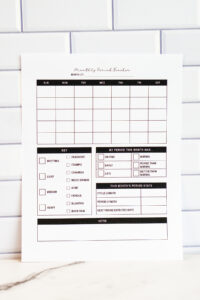 Printed monthly period tracker page shown upright in front of a white subway tile wall. The layout features an empty monthly calendar, symptom checkboxes, a section to mark period timing and quality, monthly stats, and a large notes section.
