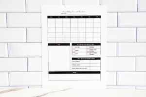Printed monthly period tracker page shown upright in front of a white subway tile wall. The layout features an empty monthly calendar, symptom checkboxes, a section to mark period timing and quality, monthly stats, and a large notes section.