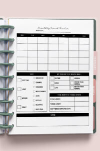Monthly period tracker printable inserted into a disc-bound planner with a blank monthly calendar, symptom checklist, period stats section, and notes area, displayed on a soft pink background.