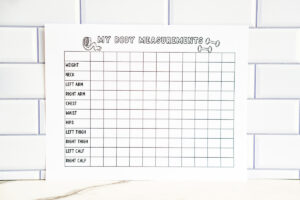 A printed &ldquo;My Body Measurements&rdquo; chart displayed upright against a white subway tile wall. The page includes a grid for tracking weight, neck, arms, chest, waist, hips, thighs, and calves, with small doodles of a tape measure and dumbbells at the top.