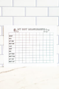 A printed &ldquo;My Body Measurements&rdquo; chart displayed against a white subway tile wall. The page includes a grid for tracking weight, neck, arms, chest, waist, hips, thighs, and calf measurements.