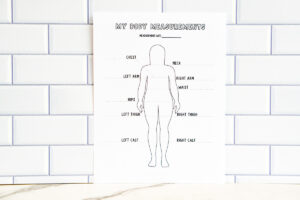 A printed full-body outline &ldquo;My Body Measurements&rdquo; sheet displayed against a subway tile wall, showing labeled lines for chest, arms, waist, hips, thighs, and calves.