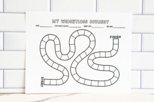 A printed game-board style &ldquo;My Weightloss Journey&rdquo; tracker displayed against a white subway tile wall. The page features a winding path of blank spaces from START to FINISH with fields for goal, each space equals, start date, and end date.