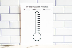A printed &ldquo;My Weightloss Journey&rdquo; thermometer-style tracker displayed upright against a white subway tile wall. The page features a large blank thermometer graphic with lines for goal, start date, and end date.