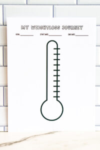 A close-up of the thermometer-style &ldquo;My Weightloss Journey&rdquo; tracker printed on white paper and displayed against a subway tile wall. The blank thermometer graphic dominates the page.