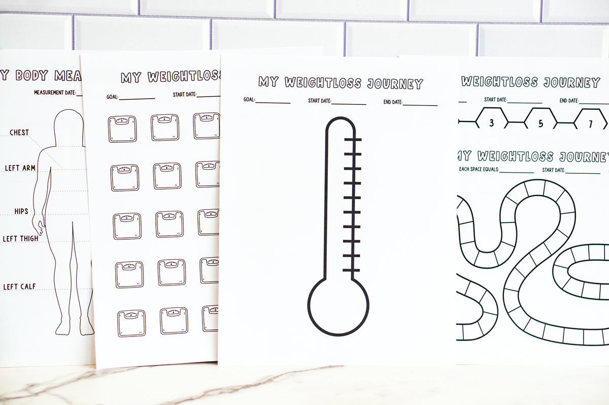 A lineup of multiple printed weight loss and measurement trackers standing side by side against a white subway tile wall, including the body outline measurement tracker, scale icon tracker, thermometer tracker, honeycomb tracker, and game-board-style tracker.