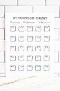 A close-up vertical view of the printed scale icon weight loss tracker against a white subway tile wall. The page includes goal, start date, and end date lines above multiple outlined scale icons.