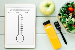 An iPad displaying a thermometer-style weight loss tracker labeled MY WEIGHTLOSS JOURNEY, placed on a mint-colored wooden surface next to a green apple, a bottle of orange juice, and a plastic container of salad with tomatoes and olives.