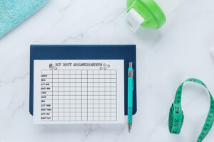 A body measurements chart titled MY BODY MEASUREMENTS sitting on a navy notebook with a teal pen, surrounded by a shaker bottle, a teal towel, and a green measuring tape on a white marble surface.