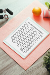 A vertical view of an iPad on a pink exercise mat displaying a hexagon-style weight loss tracker labeled MY WEIGHTLOSS JOURNEY, with dumbbells, an orange, and greenery arranged around it.