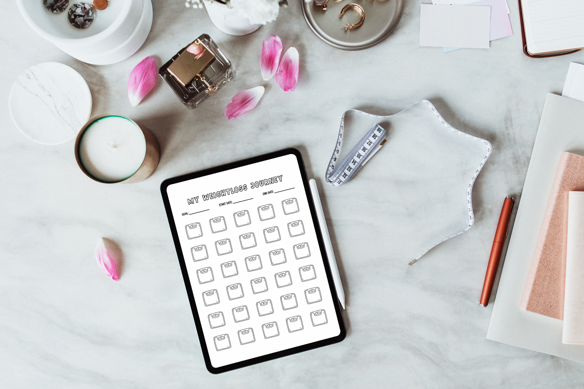 A flat-lay scene with an iPad showing a grid of scale icons on a weight loss tracker titled MY WEIGHTLOSS JOURNEY, surrounded by a candle, petals, a tape measure, jewelry dish, notebooks, and a pen on a marble surface.