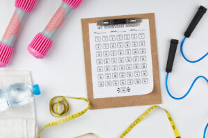 A clipboard holding a numbered scale-icon weight loss tracker labeled MY WEIGHTLOSS JOURNEY, placed among pink dumbbells, a jump rope, a water bottle, a towel, and yellow measuring tape.