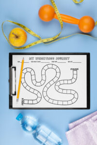 A board-game-style weight loss tracker labeled MY WEIGHTLOSS JOURNEY on a clipboard, shown with a pencil, an apple, an orange dumbbell, measuring tape, a water bottle, and a towel on a light blue background.