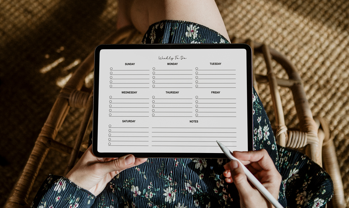 Person sitting in a wicker chair holding a tablet displaying a minimalist weekly to-do list digital planner page with sections for each day of the week and notes, using a stylus to write on the screen. The scene has a cozy, natural setting with warm lighting and a floral dress visible.