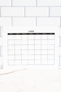 Minimalist undated June monthly calendar printed on white paper, displayed upright against a white subway tile backsplash with a marble countertop below.