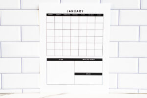 Horizontal undated January calendar page with a clean grid and planning sections below, resting against a white tile wall on a marble surface.