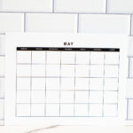 Vertical black-and-white undated May monthly calendar page propped against a white tile backsplash with a marble countertop beneath it.