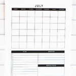 Vertical undated July calendar with grid and planning sections at the bottom, photographed upright against a white tile wall on a marble surface.