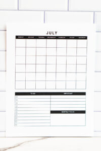 Vertical undated July calendar with grid and planning sections at the bottom, photographed upright against a white tile wall on a marble surface.
