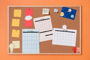 Cork bulletin board decorated with sticky notes, colorful cutouts, and three printed undated monthly calendars for October, September, and November pinned in place. The board hangs against an orange wall with a small note that reads &ldquo;Are you ready?&rdquo;