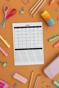 Flat lay of a printed July monthly calendar page on an orange background surrounded by colorful school and office supplies, including scissors, pencils, markers, binder clips, washi tape, and a calculator.