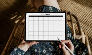 Person sitting cross-legged in a woven chair holding a digital tablet that displays an undated December monthly calendar. A stylus is held in one hand over the screen, and the person is wearing a floral dress.