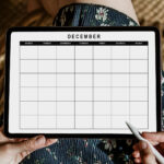 Close-up vertical view of a digital tablet displaying an undated December calendar, held by someone wearing a floral dress and holding a stylus just outside the screen.