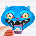 A person is holding a single KPop Demon Hunters Inspired Valentine card shaped like a blue cartoon blue Derpy from KPop Demon Hunters. The Derpy looks like a blue tiger - has bright blue skin, pointed ears, glowing yellow-to-orange eyes, and a wide open mouth with sharp white teeth. A small Valentine card sticks out of its mouth like a tongue, with one reading &ldquo;Hello Friend."
