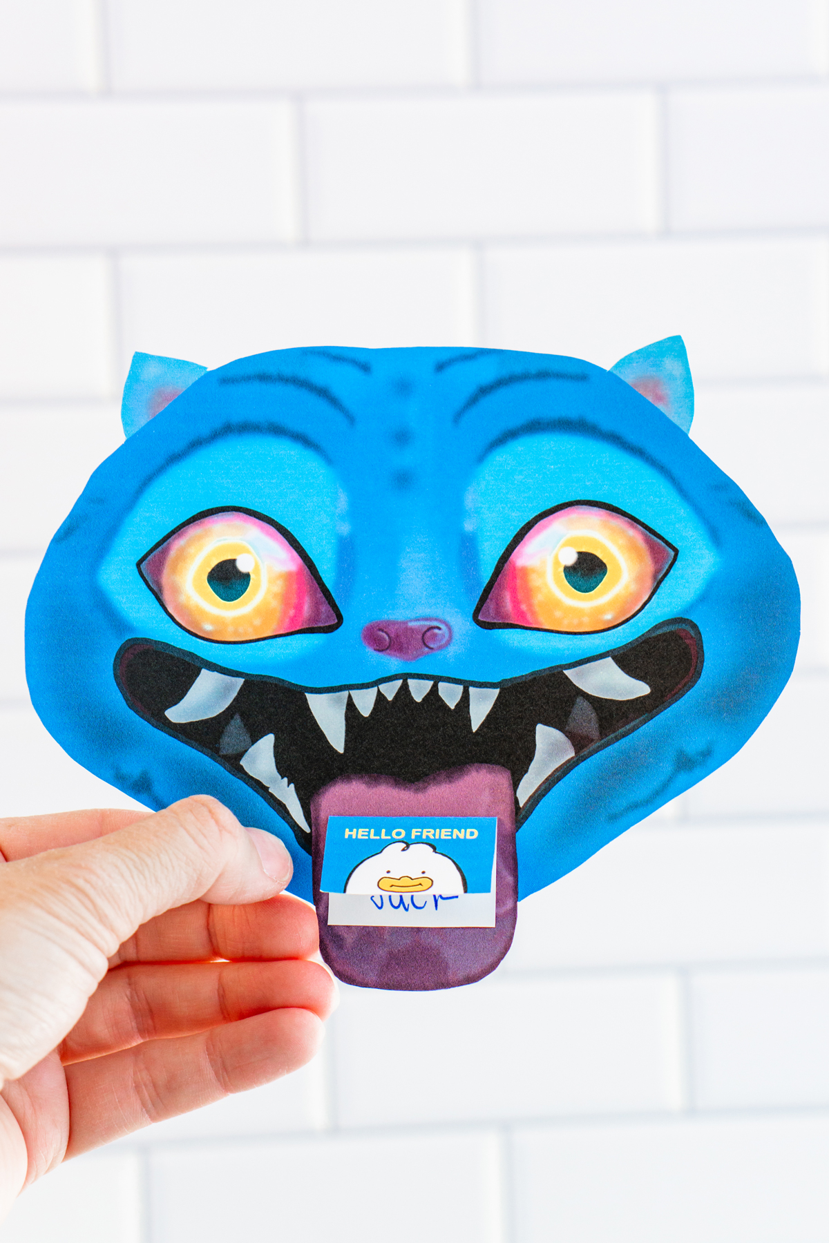 A person is holding a single KPop Demon Hunters Inspired Valentine card shaped like a blue cartoon blue Derpy from KPop Demon Hunters. The Derpy looks like a blue tiger - has bright blue skin, pointed ears, glowing yellow-to-orange eyes, and a wide open mouth with sharp white teeth. A small Valentine card sticks out of its mouth like a tongue, with one reading &ldquo;Hello Friend."