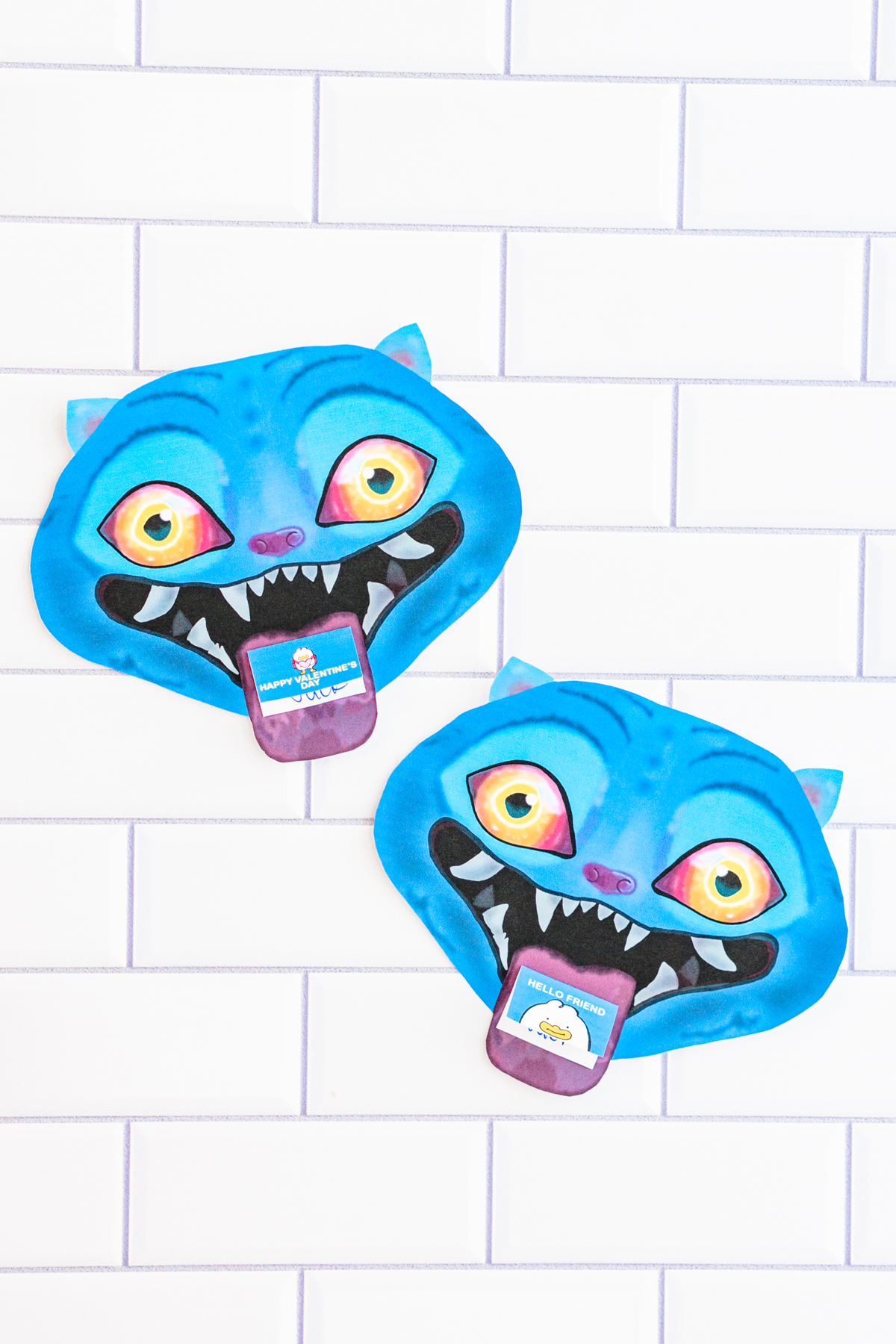 Two finished Valentine cards shaped like a blue cartoon blue Derpy from KPop Demon Hunters. They are placed on a white tile surface. Each demon has bright blue skin, pointed ears, glowing yellow-to-orange eyes, and a wide open mouth with sharp white teeth. A small Valentine card sticks out of each demon's mouth like a tongue, with one reading 