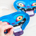 A close-up shows an adult hand holding a small white handwritten note against the purple tongue of Derpy, a blue demon character from KPop Demon Hunters. The note reads &ldquo;Your friend, Jack&rdquo; in blue ink. Derpy&rsquo;s bright yellow-and-pink eyes, sharp white teeth, and playful expression are visible in the background.