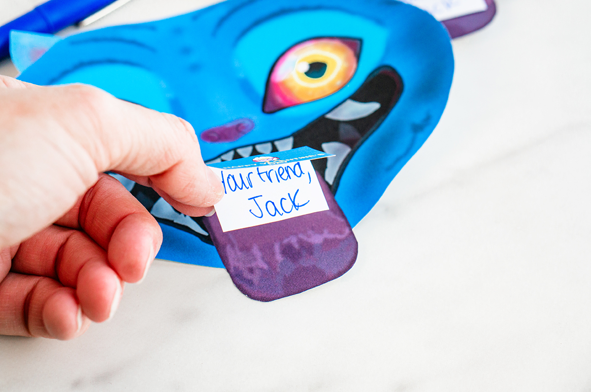 A close-up shows an adult hand holding a small white handwritten note against the purple tongue of Derpy, a blue demon character from KPop Demon Hunters. The note reads &ldquo;Your friend, Jack&rdquo; in blue ink. Derpy&rsquo;s bright yellow-and-pink eyes, sharp white teeth, and playful expression are visible in the background.