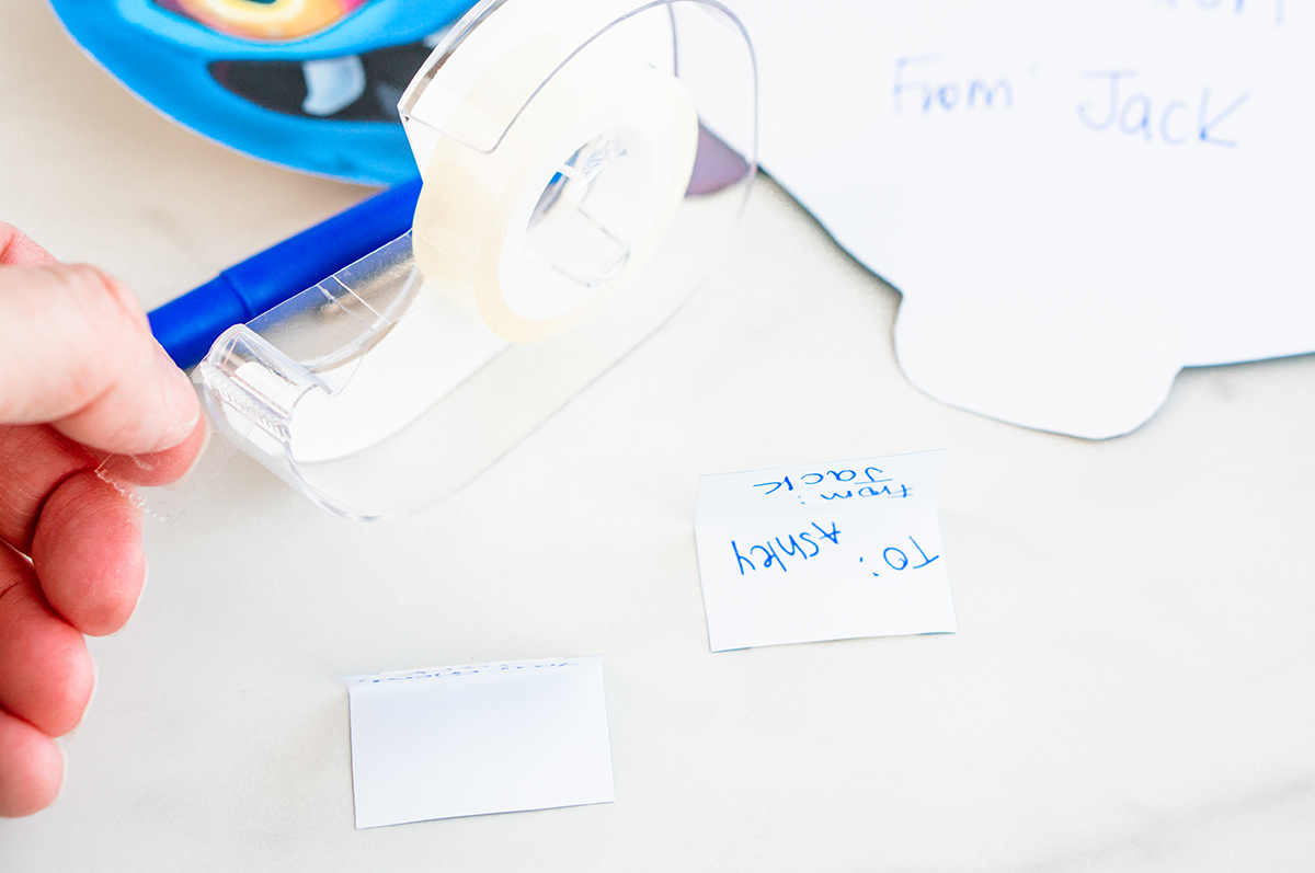 Another close-up focuses on the tape dispenser and a small white folded note lying flat on the table. A handwritten note reading &ldquo;To: Ashley From: Jack&rdquo; is nearby, reinforcing the step of preparing name labels before attaching them.