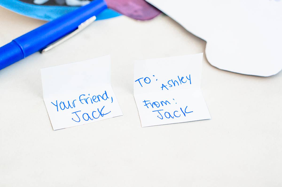 A flat lay shows two small handwritten white notes side by side. One reads &ldquo;Your friend, Jack,&rdquo; and the other reads &ldquo;To: Ashley From: Jack,&rdquo; written in blue ink. A blue pen and part of the Derpy Valentine cutout are visible nearby.