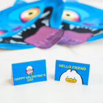 A slightly blurred close-up shows two folded blue Valentine message cards standing upright on a white surface. One reads &ldquo;Happy Valentine&rsquo;s Day&rdquo; with a cartoon duck holding hearts, and the other reads &ldquo;Hello Friend&rdquo; above a duck face illustration. Two Derpy Valentine cutouts appear softly blurred in the background.