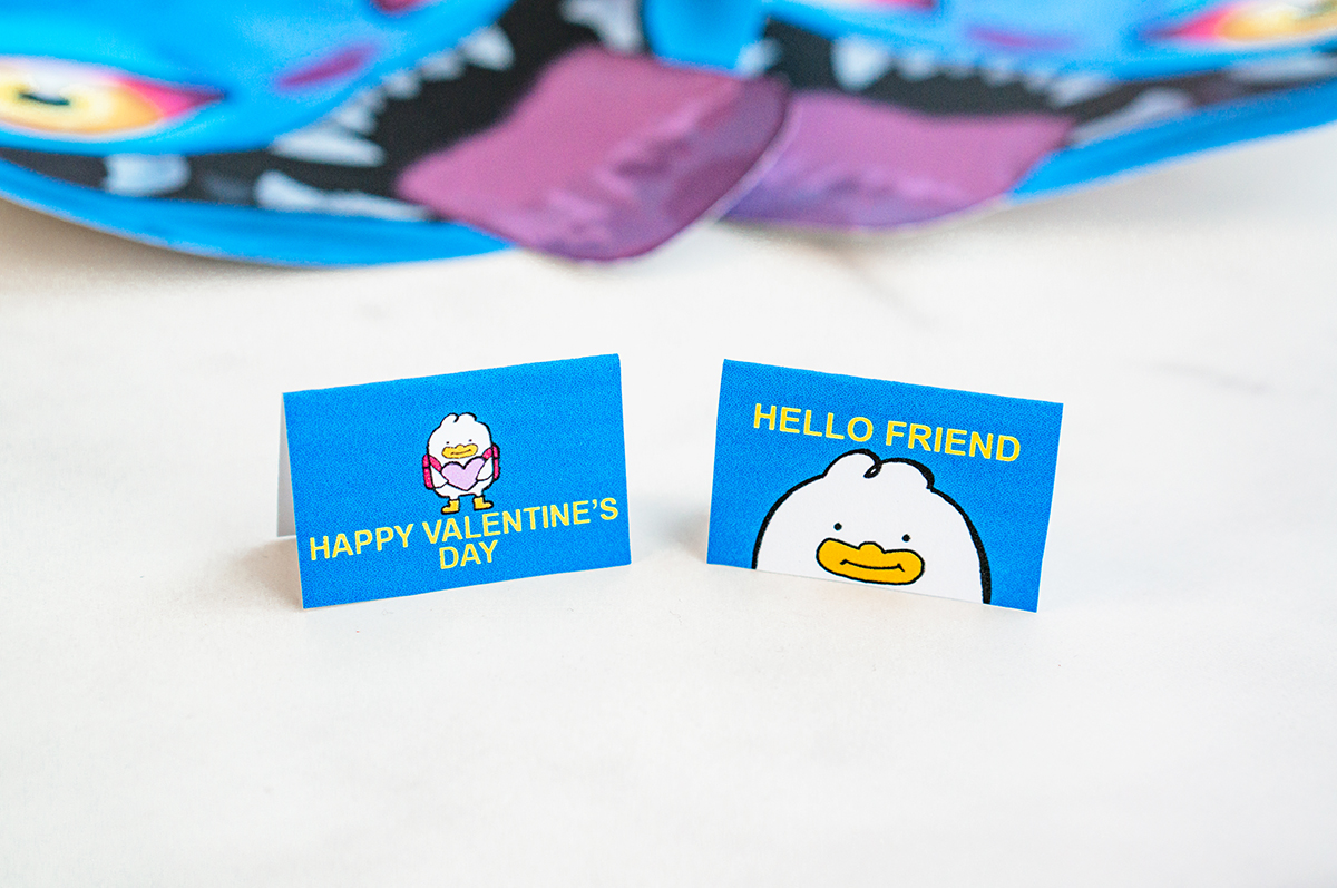A slightly blurred close-up shows two folded blue Valentine message cards standing upright on a white surface. One reads &ldquo;Happy Valentine&rsquo;s Day&rdquo; with a cartoon duck holding hearts, and the other reads &ldquo;Hello Friend&rdquo; above a duck face illustration. Two Derpy Valentine cutouts appear softly blurred in the background.