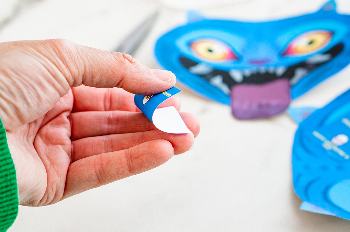 A close-up shows an adult hand holding a folded blue message card as it is gently curved, demonstrating how the card will be folded to fit onto Derpy&rsquo;s tongue. Finished Derpy Valentine cutouts are visible in the background on the white surface.
