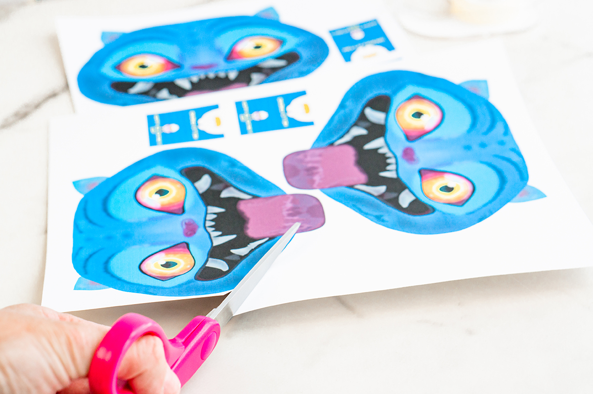 A close-up action shot shows pink-handled scissors cutting along the edge of a printed sheet containing multiple blue Derpy Valentine faces and small blue message cards. The scissors are mid-cut, demonstrating how the printable Valentines are separated from the page.