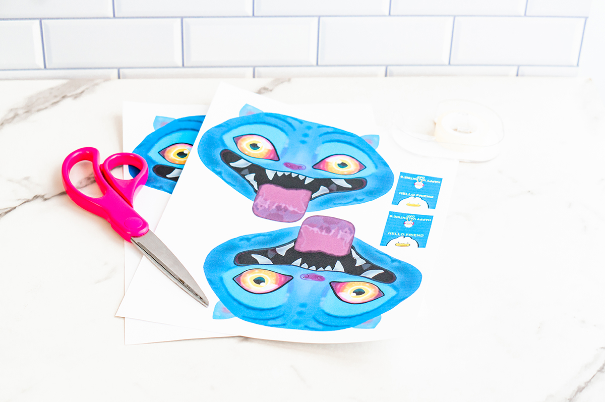 Two printed sheets of blue cartoon Derpy from KPop Demon Hunters Valentine designs lie slightly stacked on a white marble countertop with a white subway tile wall in the background. Each sheet shows a bright blue Derpy face with glowing yellow-orange eyes, pointed ears, and a wide open mouth with sharp white teeth and a purple tongue. Pink-handled scissors rest to the left, and small rectangular Valentine message cards are printed on the right side of the page, showing how the Valentine will be assembled.